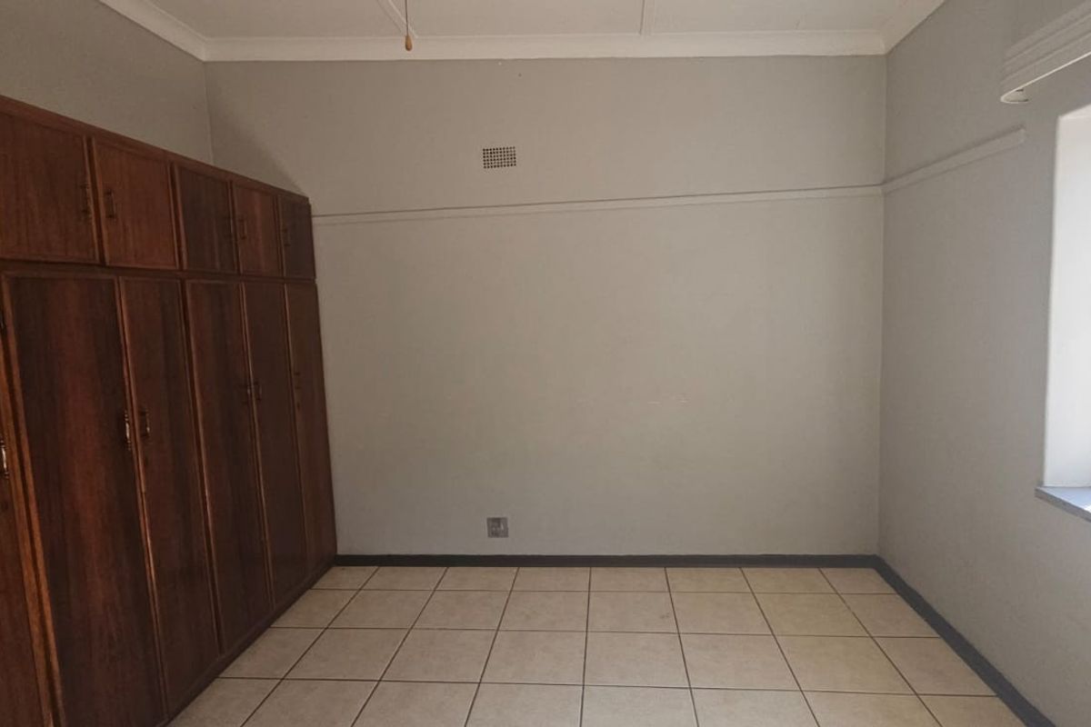 A spacious, empty bedroom featuring tiled flooring, built-in wooden cupboards, and a window fitted with security bars.