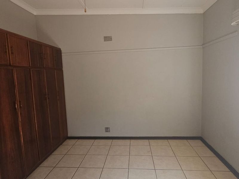 A spacious, empty bedroom featuring tiled flooring, built-in wooden cupboards, and a window fitted with security bars.