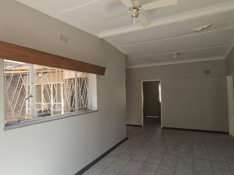 An empty living area featuring tiled flooring, a ceiling fan, and security bars on the window, with visible damage to the ceiling plaster.