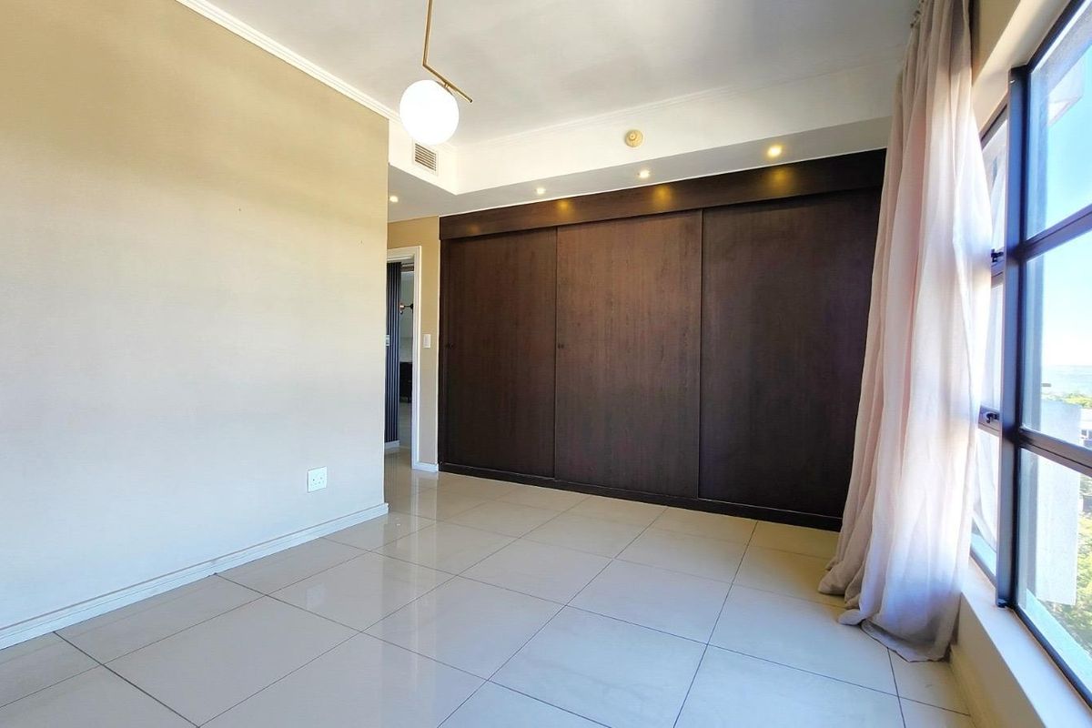 A bright, empty bedroom featuring large windows, tiled flooring, and a spacious built-in sliding door wardrobe with integrated recessed lighting.