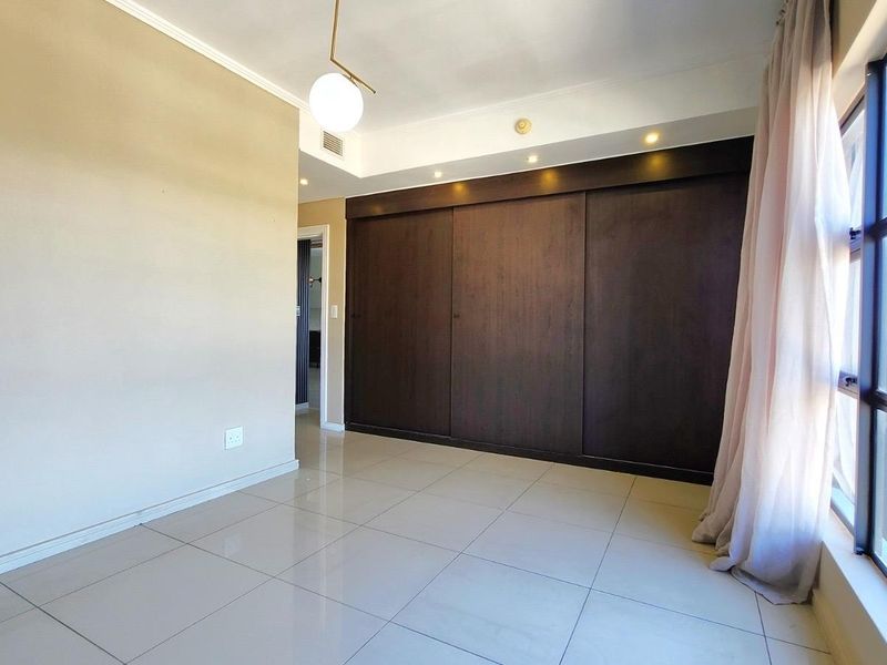 A bright, empty bedroom featuring large windows, tiled flooring, and a spacious built-in sliding door wardrobe with integrated recessed lighting.
