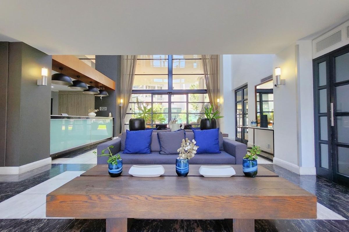 Spacious living area featuring double volume ceilings, large glass windows for natural light, and modern tiled flooring in a well-maintained apartment setting.
