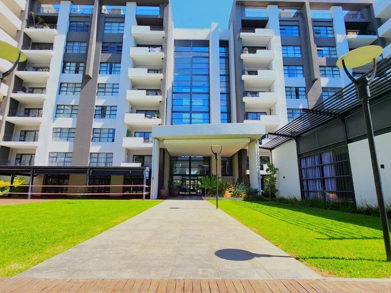 Modern multi-story apartment building exterior featuring a central glass facade, covered entrance portico, and well-maintained landscaped grounds.