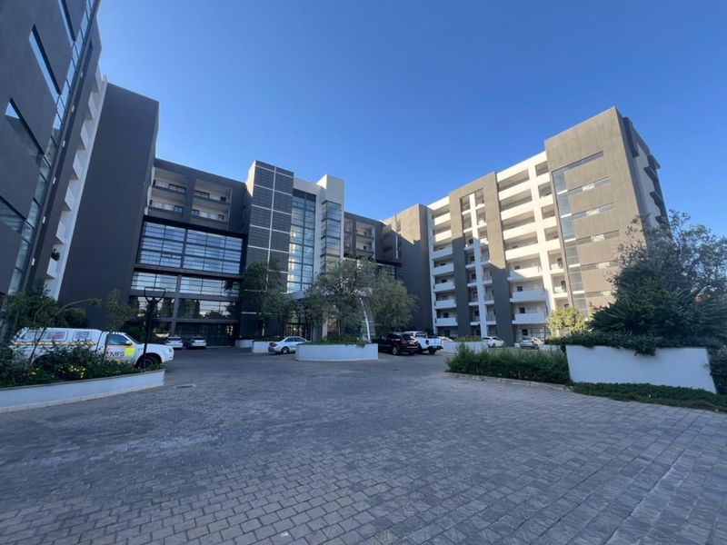 A modern multi-story apartment complex featuring a paved entrance courtyard, contemporary facade design with glass elements, and established landscaped garden beds.
