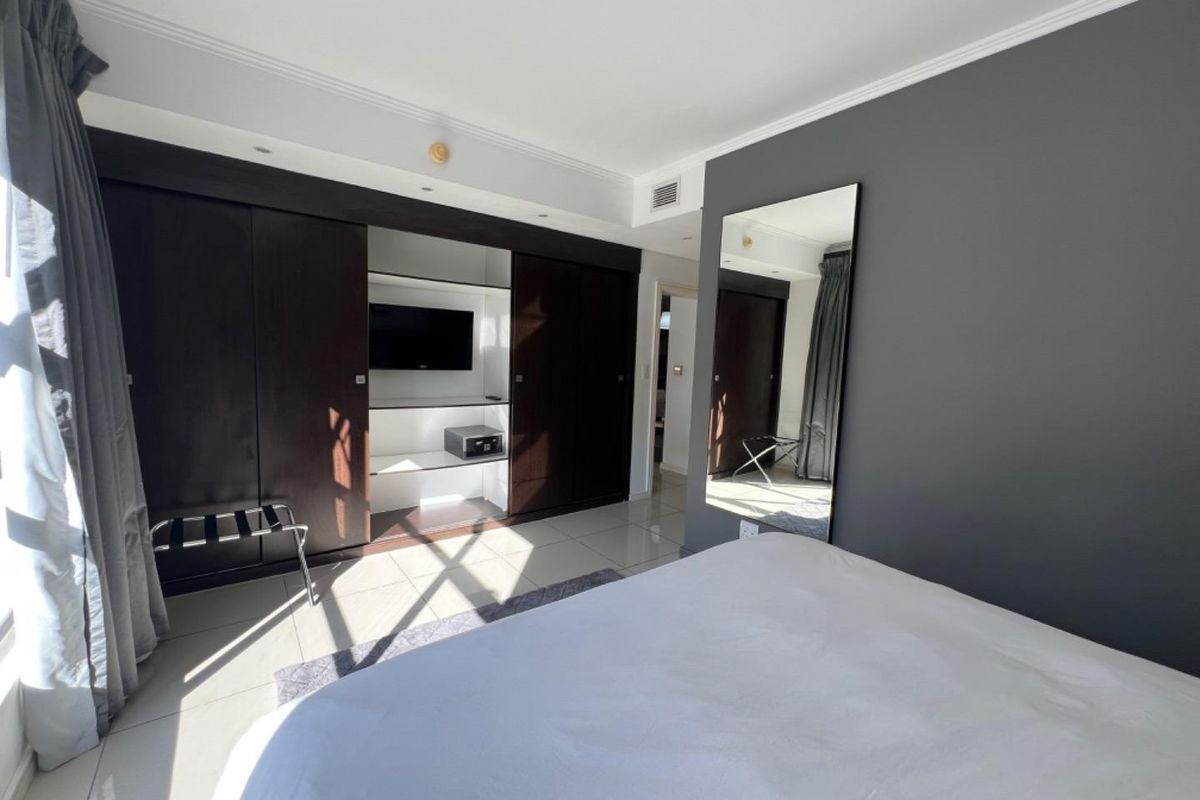 A modern bedroom featuring dark built-in cupboards with an integrated media unit, tiled flooring, and a large wall-mounted mirror.