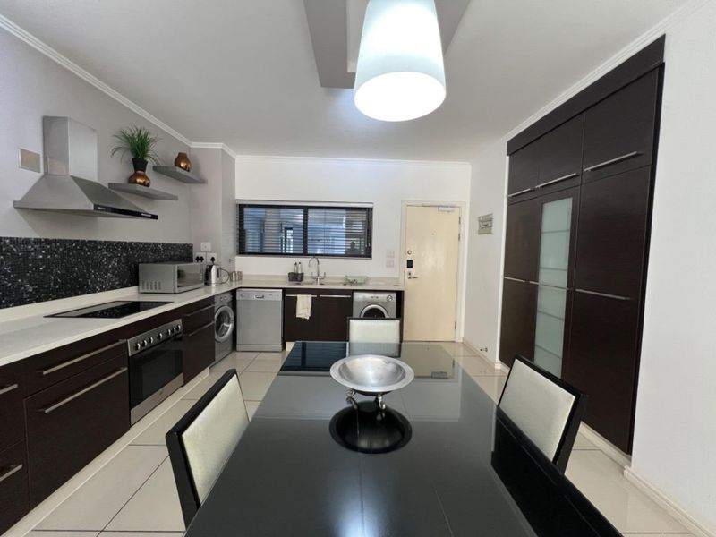 Modern open-plan kitchen featuring dark cabinetry, integrated appliances, mosaic backsplash, and tiled flooring, seamlessly flowing into the dining area.