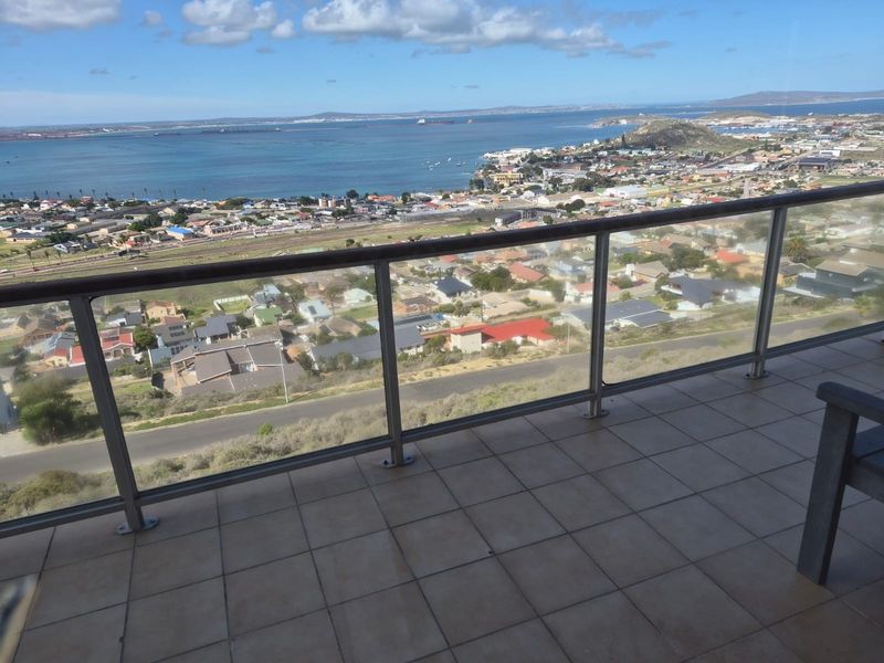 A spacious balcony featuring a modern glass balustrade and tiled flooring, offering expansive panoramic views of the coastal town and bay.