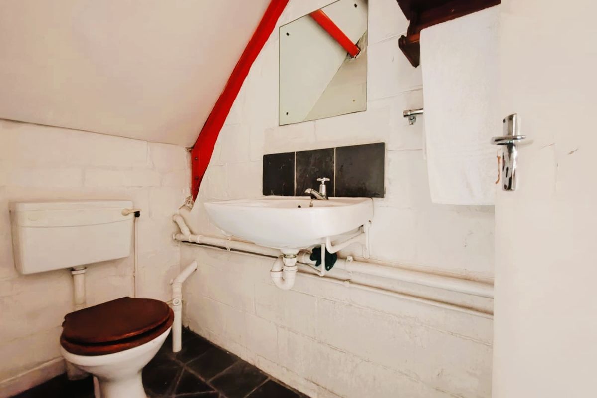 A compact bathroom featuring a wall-mounted basin, toilet, and exposed plumbing, set within a space with sloped ceilings and painted brick walls.