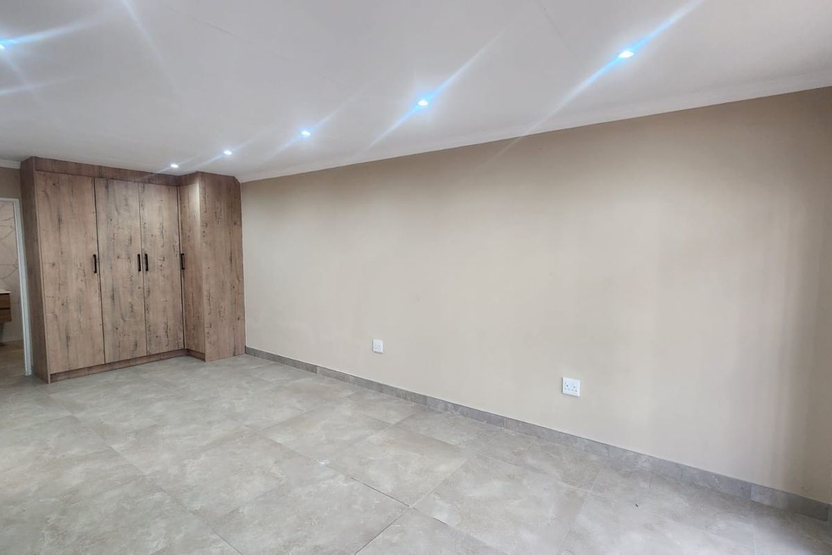 A bright, modern bedroom featuring neutral walls, large format floor tiles, and integrated wood-finish built-in cupboards with recessed ceiling lighting.