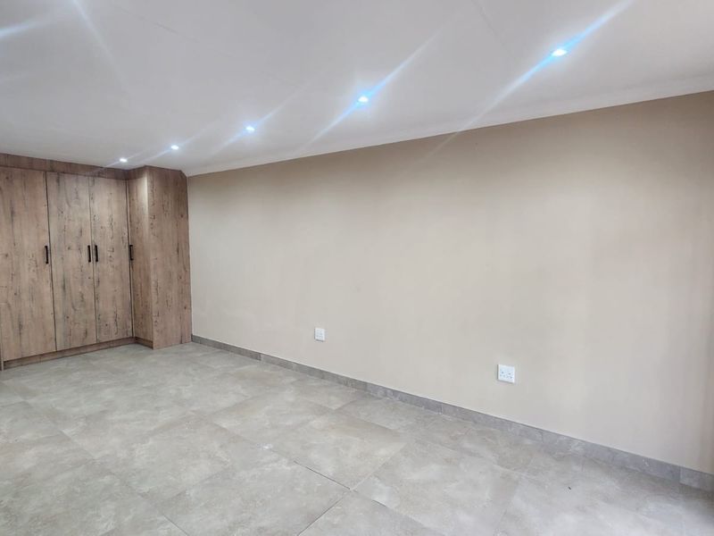 A bright, modern bedroom featuring neutral walls, large format floor tiles, and integrated wood-finish built-in cupboards with recessed ceiling lighting.