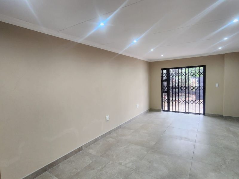 A bright, empty living area featuring neutral tiled flooring, recessed downlighting, and a secure metal gate leading to the exterior.