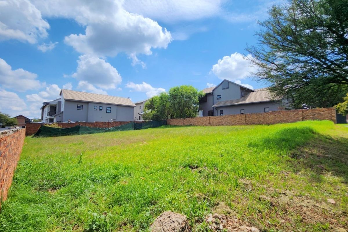 A vacant residential plot featuring a grassy landscape, enclosed by brick boundary walls and green mesh fencing, situated in a developed residential area.
