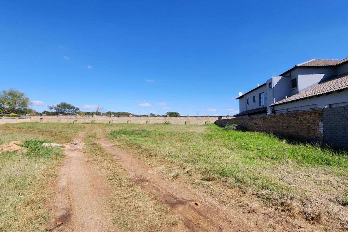 A vacant residential plot featuring a dirt track, grass coverage, and a perimeter boundary wall adjacent to an existing residential structure.