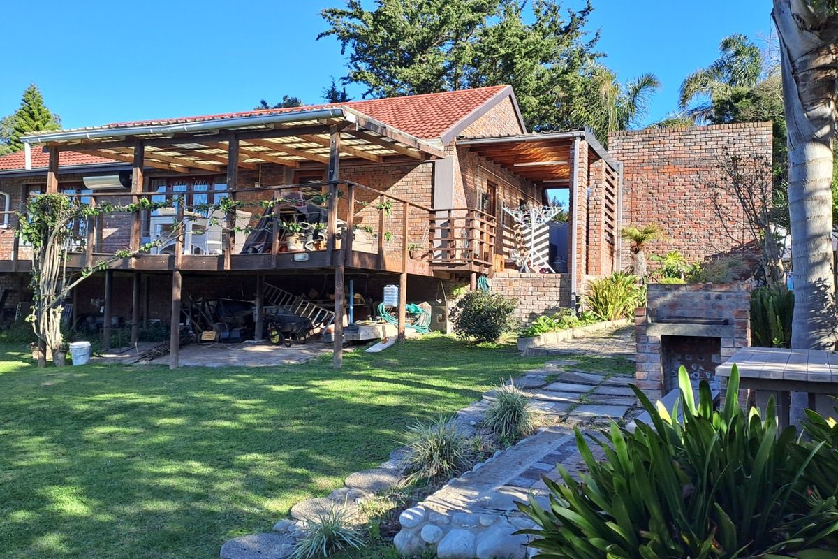 Rear view of the house featuring a raised timber deck, covered patio area, built-in brick braai, and a well-maintained garden with stone pathways.
