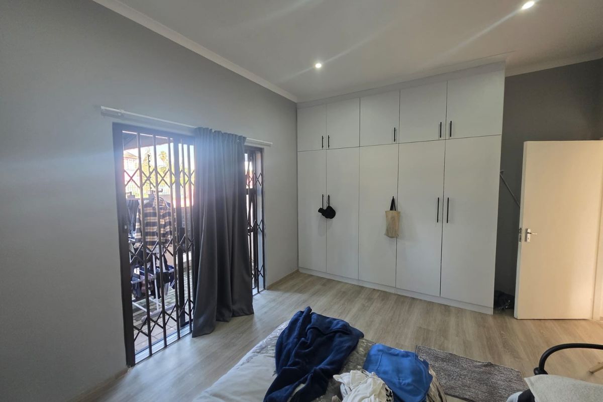 A well-maintained bedroom featuring modern built-in cupboards, light laminate flooring, recessed downlighting, and a sliding door leading to an outdoor area with security gate.