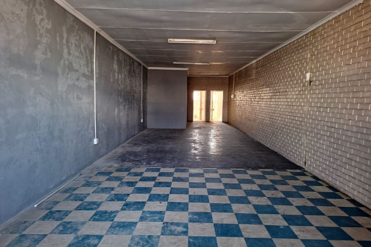 A vacant, open-plan commercial space featuring a mix of concrete and checkered flooring, exposed brickwork, and functional overhead lighting.