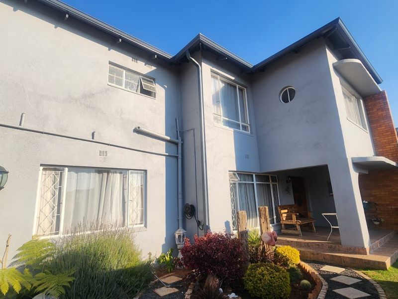 Rear view of a double-story house featuring a tiled patio, plastered walls with some staining, and established garden beds.