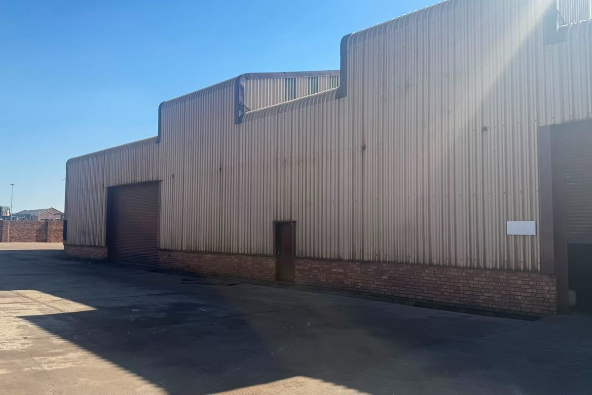Industrial warehouse exterior featuring corrugated metal cladding, brick base, and multiple roller shutter doors with an expansive concrete yard area.
