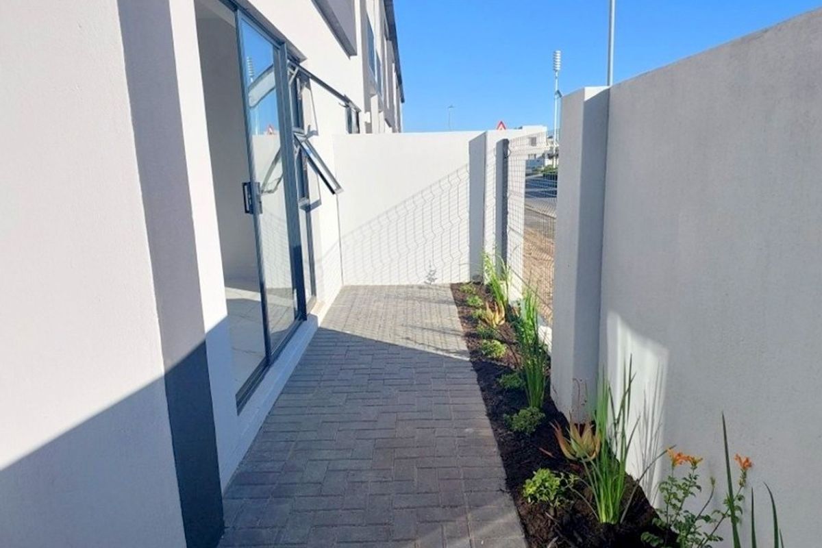 A clean, modern exterior rear walkway featuring paved flooring, a boundary wall, and direct access to the interior via a sliding glass door.