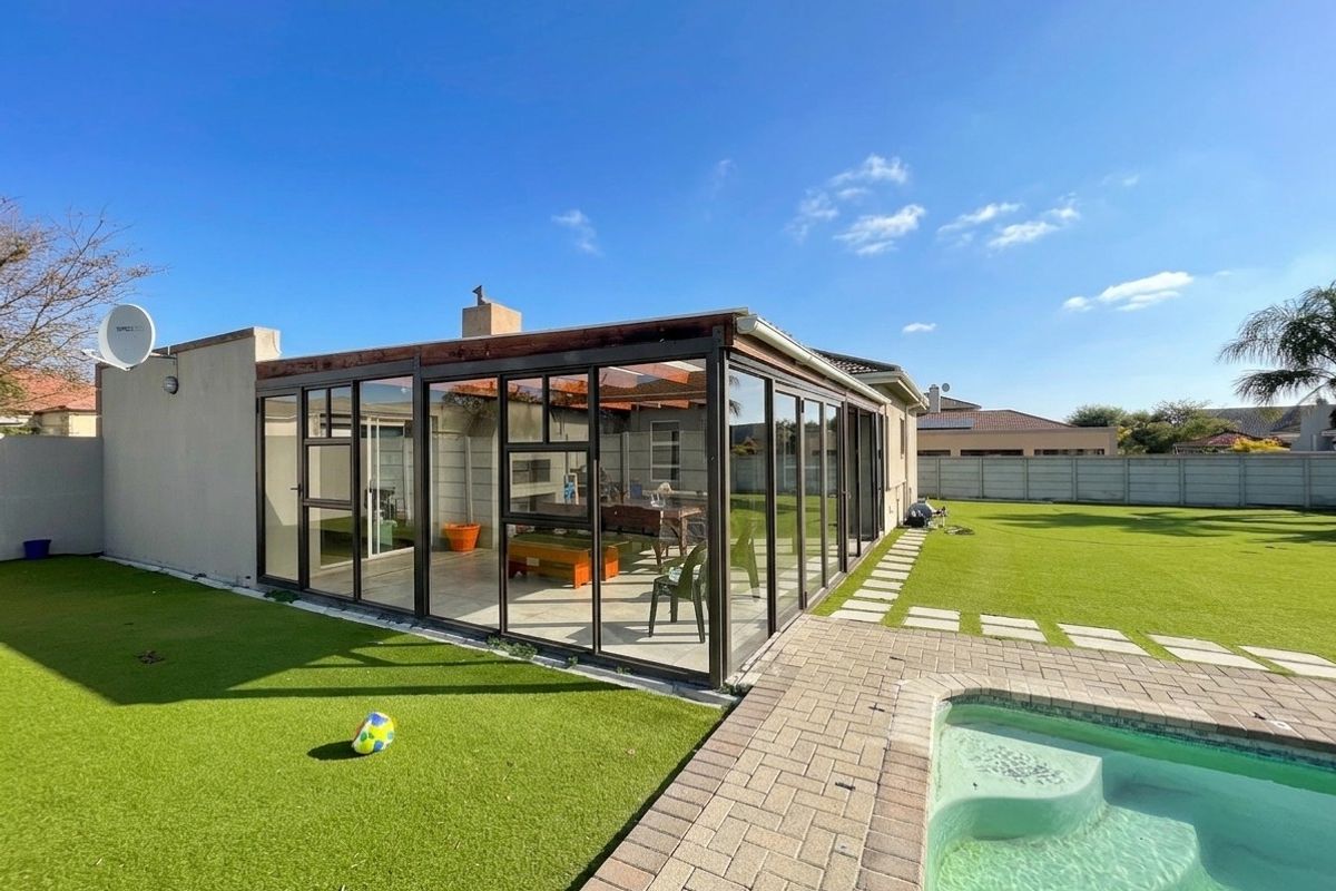 Rear view of the property featuring an enclosed glass patio, a swimming pool with paver surround, and a well-maintained artificial lawn.