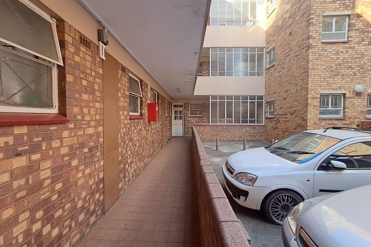 Exterior view of an apartment building walkway featuring brick construction, tiled flooring, and steel-framed windows overlooking a parking area.