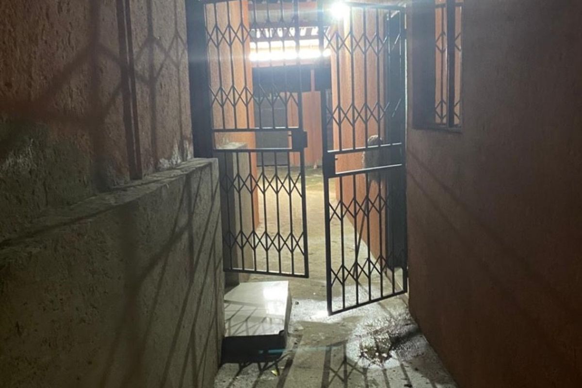 A narrow exterior side passage at night featuring a metal security gate and concrete flooring between two building walls.