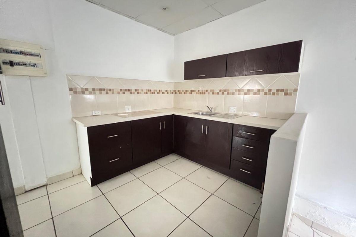 A compact, empty kitchen featuring dark wood cabinetry, laminate countertops, a stainless steel sink, and tiled backsplash, suitable for a commercial office or small unit.
