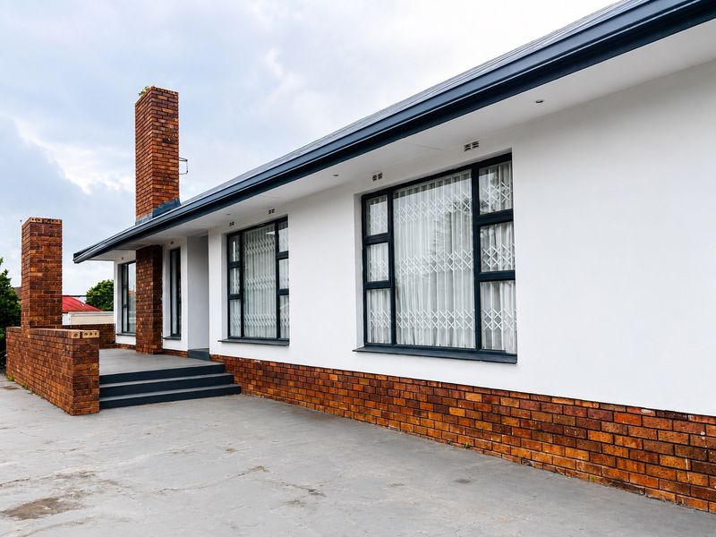 A well-maintained exterior view of the guest house featuring a clean white facade, brick accents, prominent chimneys, and secure aluminium-framed windows.