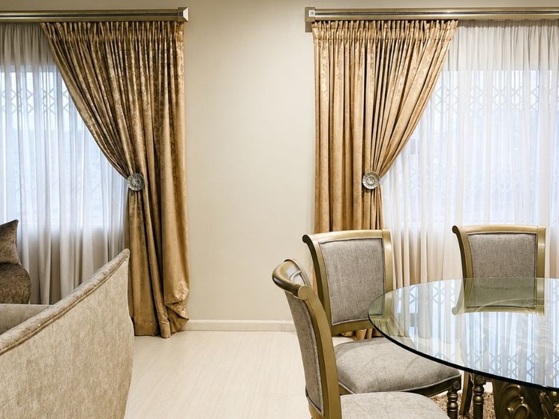 A well-maintained dining area featuring neutral walls, laminate flooring, recessed lighting, and large windows fitted with decorative curtains and security gates.