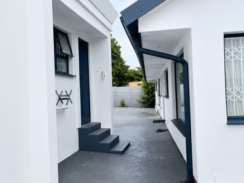 A clean exterior view of the guest house rear pathway, featuring modern white facades, dark trim, and paved access between building sections.