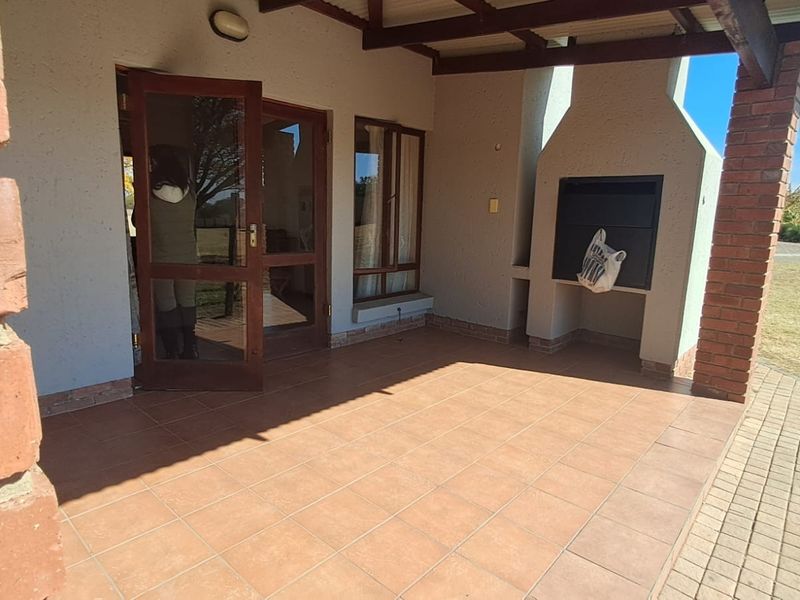 Covered patio featuring tiled flooring, a built-in braai, and timber-framed glass doors leading into the home, overlooking the garden area.