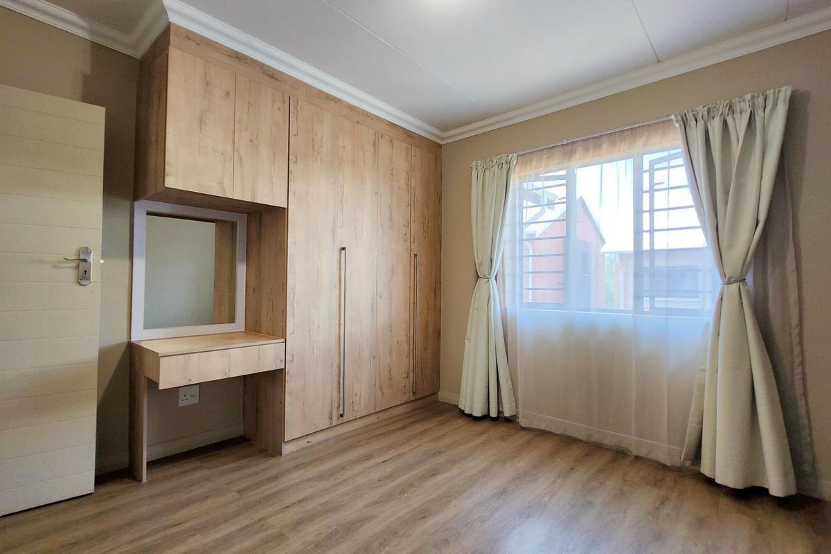 A bright bedroom featuring light wood-look laminate flooring, extensive built-in cupboards with an integrated vanity, and a window fitted with security bars.