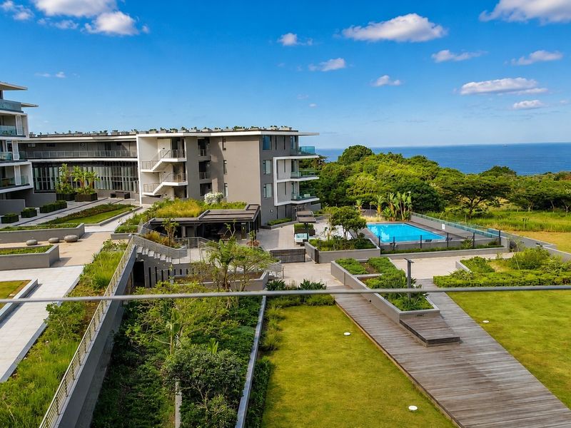 Elevated view of the apartment complex grounds featuring landscaped gardens, timber decking, paved pathways, and a communal swimming pool with ocean views.