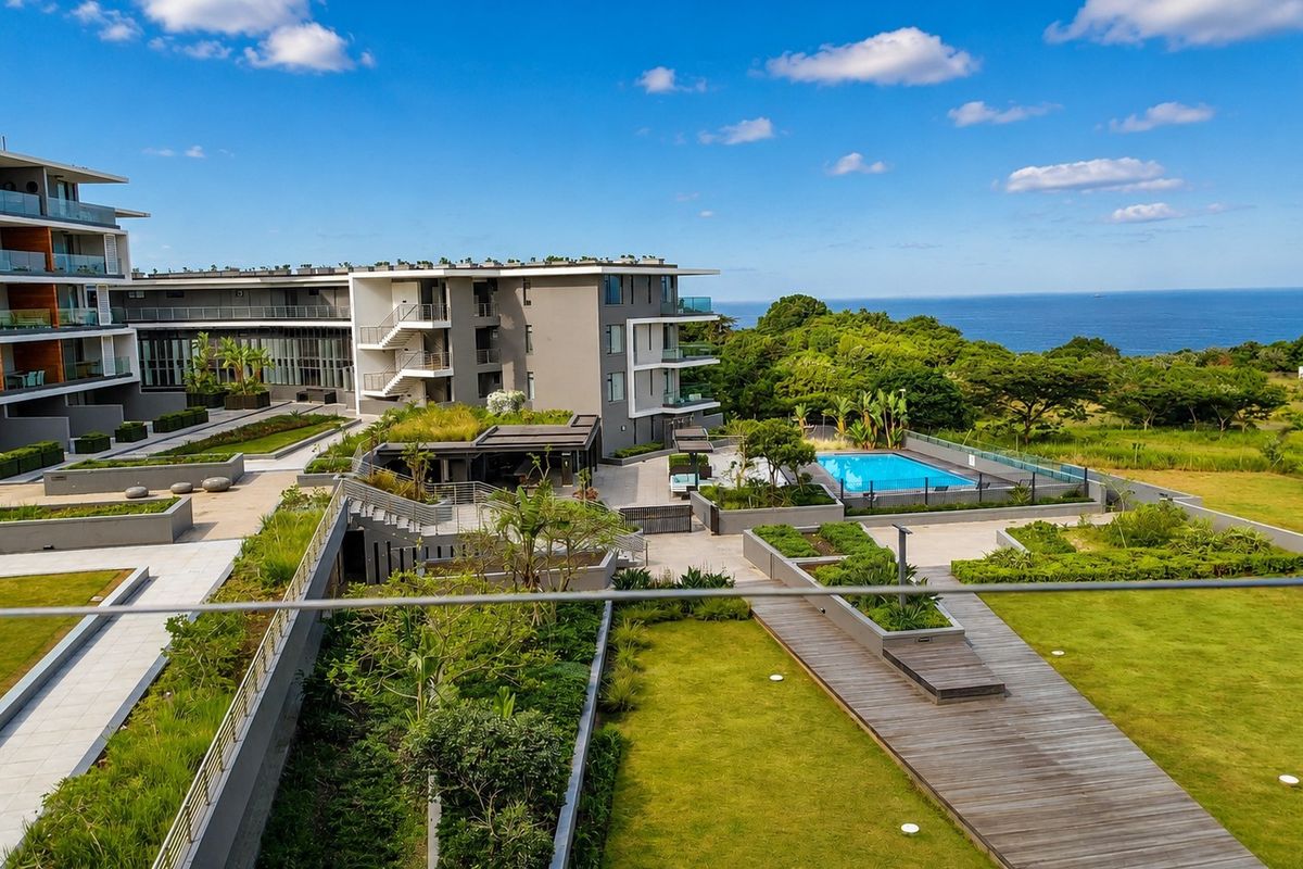 Elevated view of the apartment complex grounds featuring landscaped gardens, timber decking, paved pathways, and a communal swimming pool with ocean views.