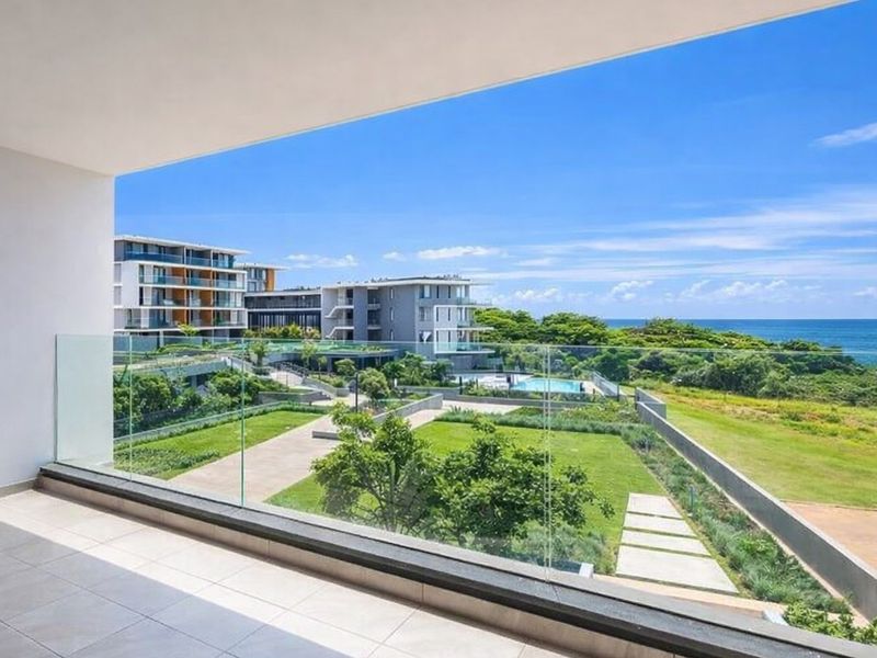 A spacious balcony featuring a modern glass balustrade, offering expansive views over the landscaped estate grounds and the ocean beyond.