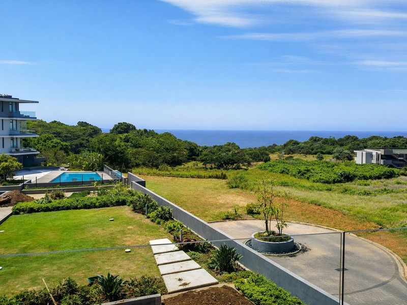 Elevated coastal view overlooking a landscaped garden, swimming pool, and paved driveway with the ocean visible in the distance.