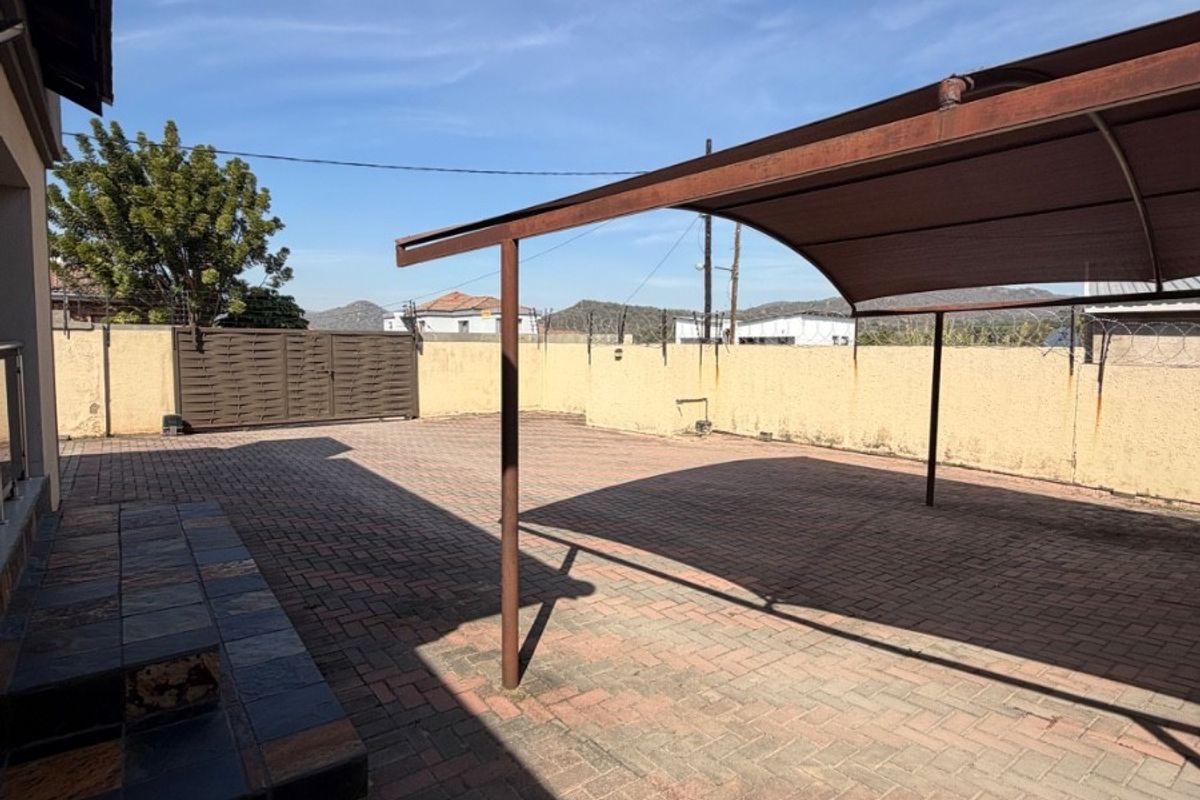 A spacious paved rear courtyard featuring a metal carport structure, perimeter walling with security fencing, and tiled steps leading to the main dwelling.