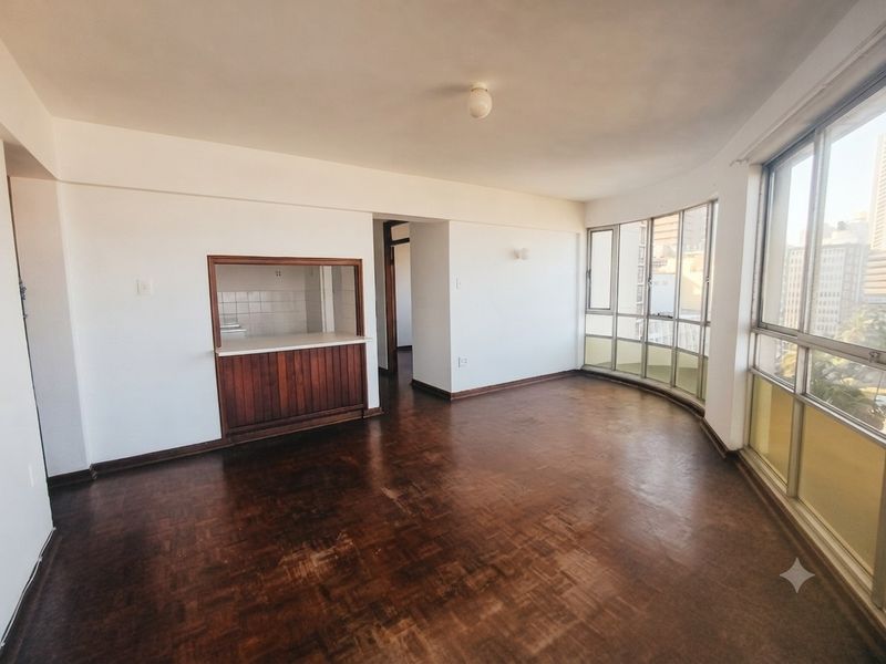 Spacious living area featuring original parquet flooring, large windows with city views, and a convenient serving hatch connecting to the kitchen area.