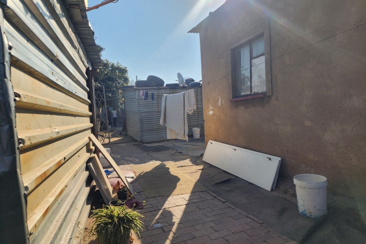 Rear exterior view showing a paved walkway between a corrugated metal structure and a plastered masonry building.