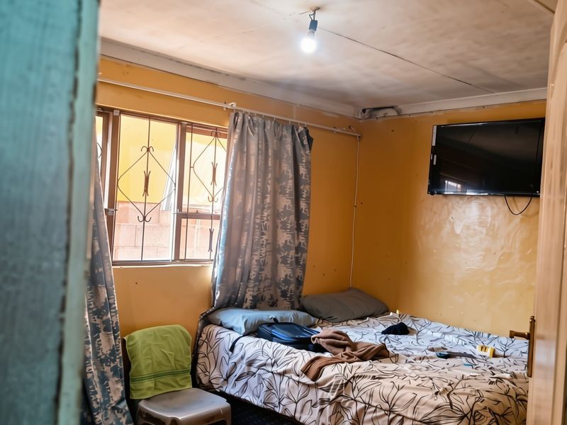 A compact bedroom featuring tiled flooring, security bars on the window, and a wall-mounted television, requiring cosmetic updates to improve the overall finish.