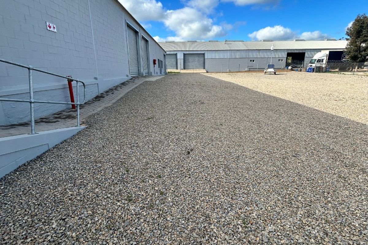 Spacious gravel yard area providing access to commercial warehouse units with roller shutter doors and metal cladding exterior.