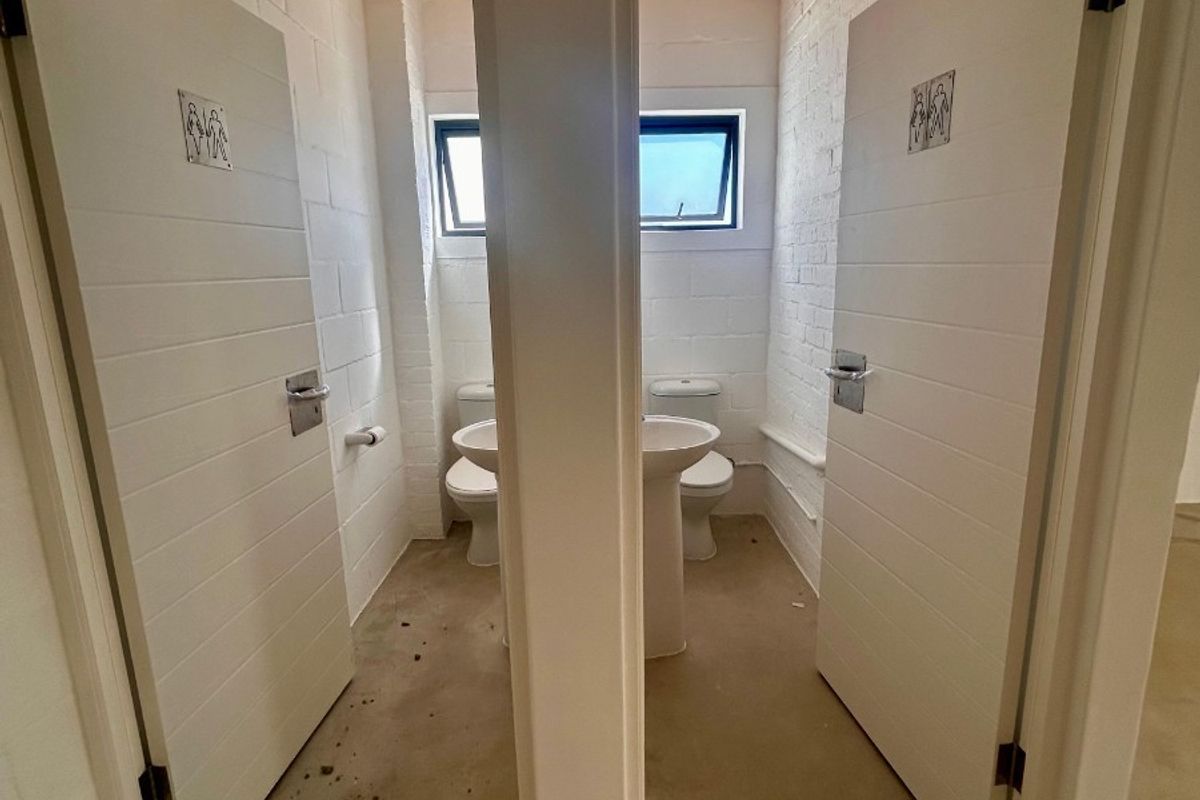 Two separate restroom cubicles featuring pedestal sinks, toilets, painted brick walls, and exposed timber ceilings, requiring cosmetic updates to the flooring and finishes.