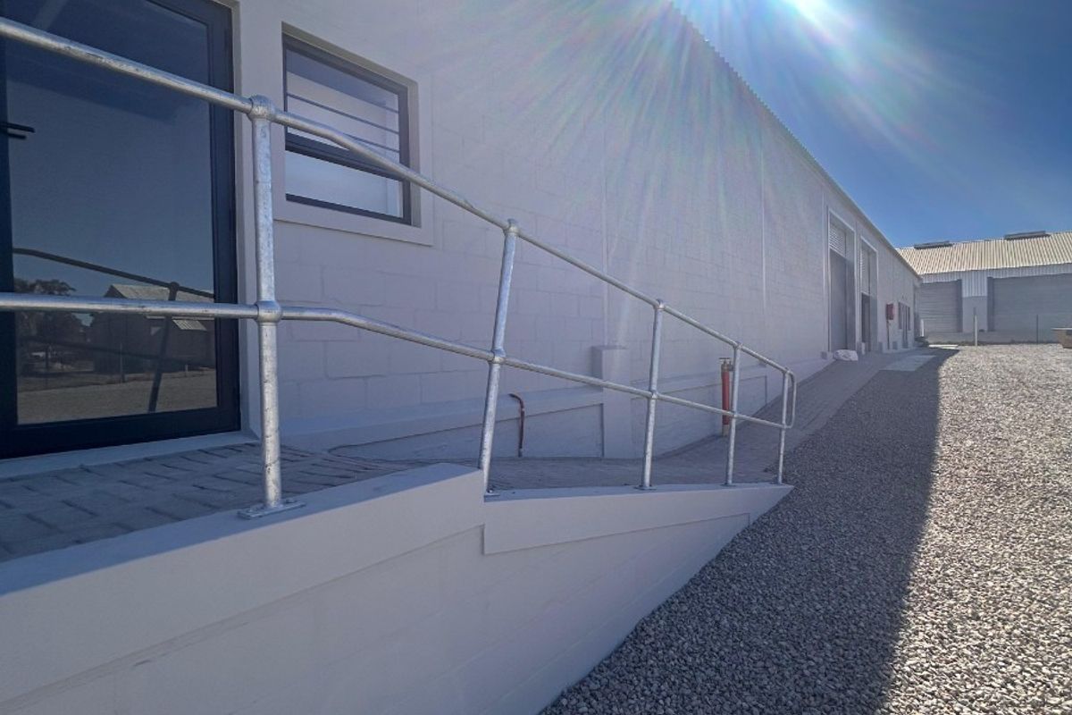 Exterior view of a commercial building featuring a concrete access ramp with a galvanized steel handrail and gravel driveway.