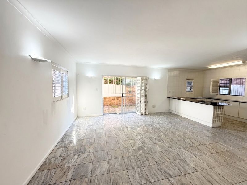 Spacious open-plan living area featuring tiled floors, a functional kitchen with built-in cabinetry, and direct access to the rear exterior through a security gate.