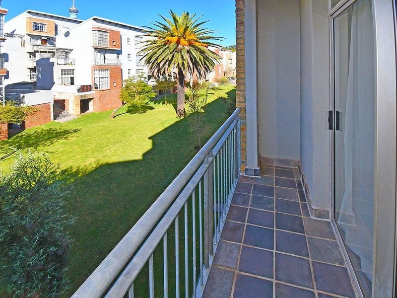 A private balcony featuring tiled flooring and a metal balustrade, offering an elevated view over the complex's landscaped garden and surrounding apartment buildings.