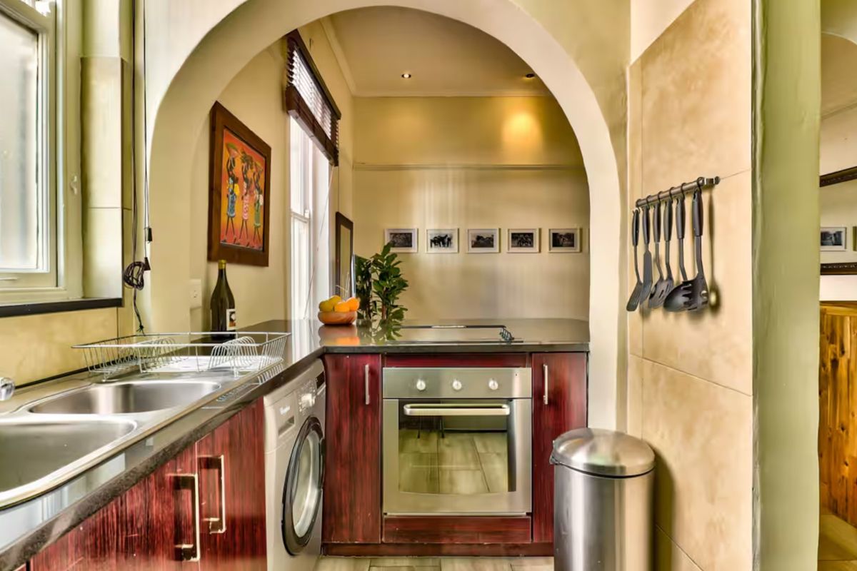 A compact kitchen featuring dark wood-finish cabinetry, a built-in oven, stainless steel sink, and a distinctive architectural archway connecting to the adjacent living space.