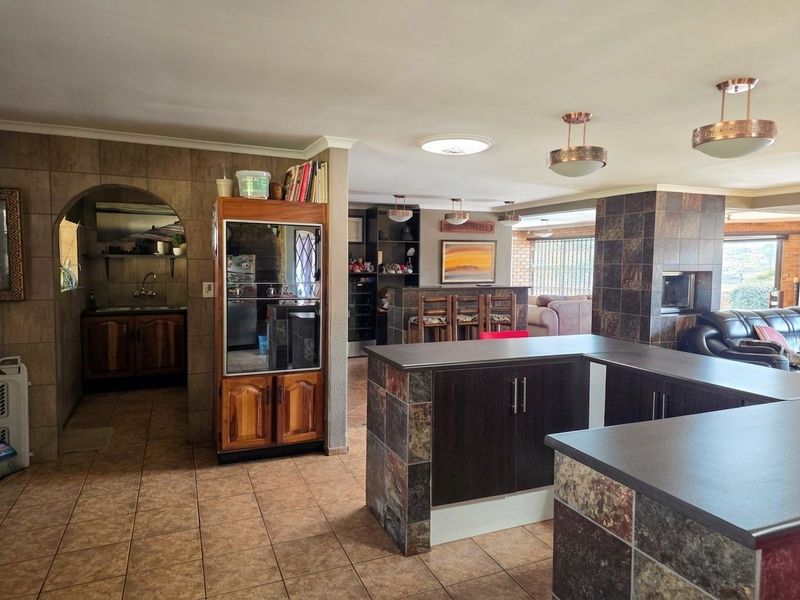 Spacious open-plan living area featuring a stone-clad kitchen island, built-in fireplace, and tiled flooring, connecting seamlessly to a secondary kitchen or scullery area.