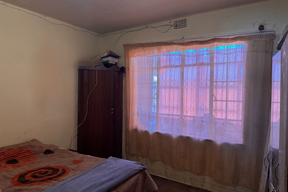 A bedroom featuring parquet flooring, a window with security bars, and visible surface wiring, requiring cosmetic updates to improve its overall condition.