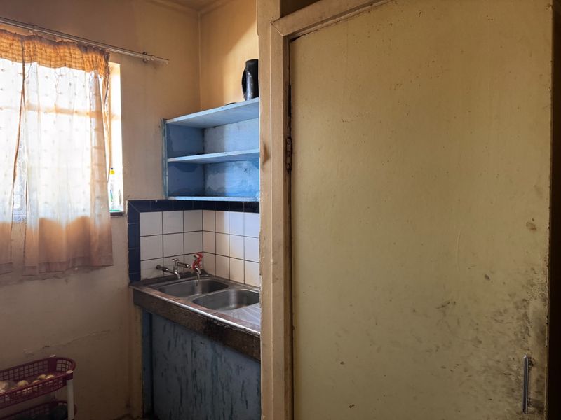 A compact scullery area featuring a double basin sink, tiled splashback, and open shelving, requiring cosmetic updates and refurbishment.