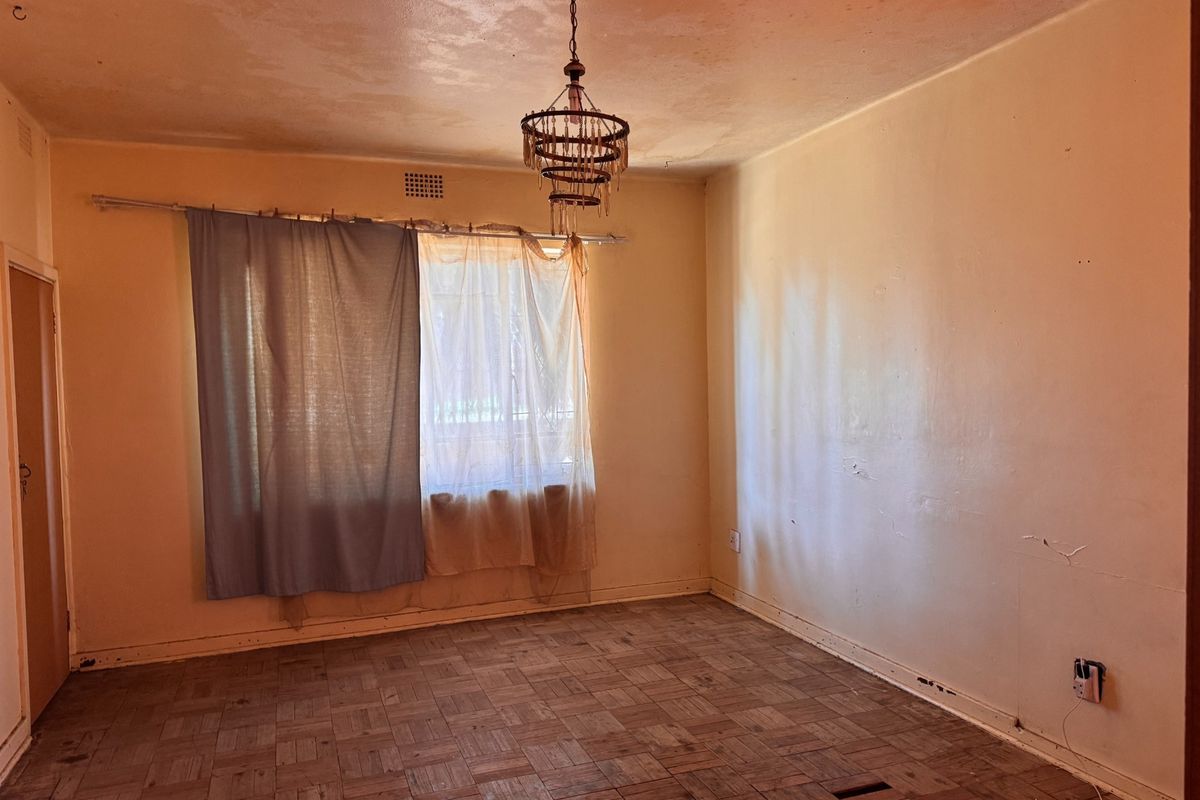 An empty living room featuring original parquet flooring, a central light fixture, and large windows, requiring cosmetic updates and floor restoration.